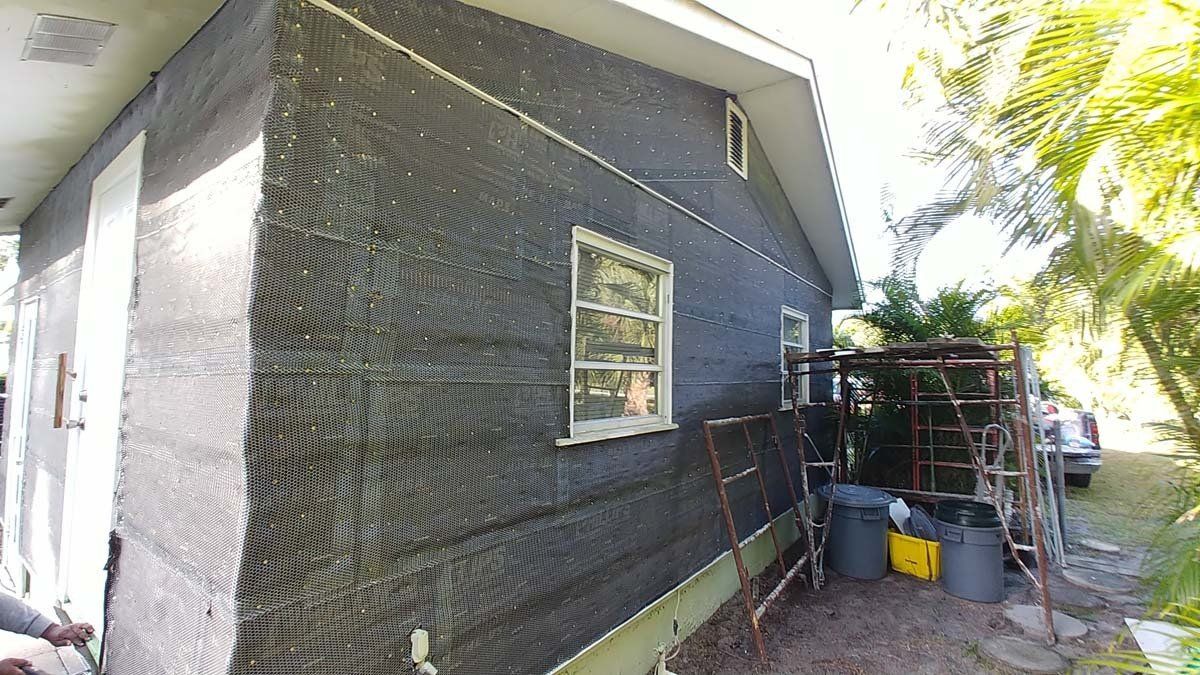 Side of a house with dark siding, a small window, and outdoor storage items along a narrow path.