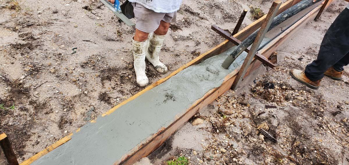 Workers carrying a long, wet concrete slab with orange edging at a muddy construction site