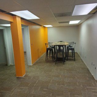 Empty office room with orange wall, white ceiling lights, carpet, and a high table with stools in the back.