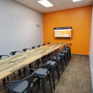 Conference room with long wooden table, black chairs, and orange accent wall with wall-mounted screen.