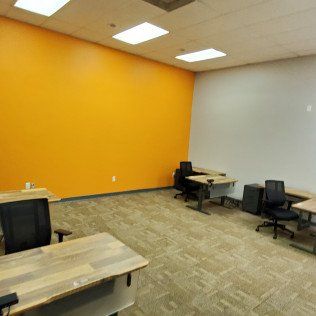 Empty office room with orange accent wall, gray carpet, desks, and chairs under ceiling lights