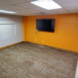 Empty office room with orange walls, wall-mounted TV, carpeted floor, and a window with blinds.