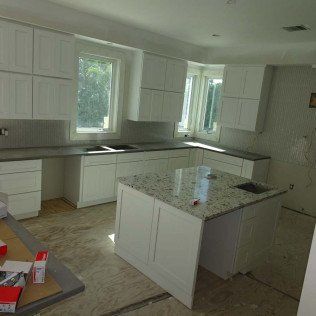 Bright unfinished kitchen with white cabinets, granite island, and light wood floor under renovation