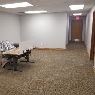 Empty office hallway with gray walls, carpeted floor, a bench, and a closed door at the end