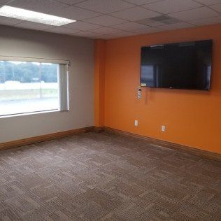 Empty office waiting room with orange accent wall, wall-mounted TV, window, and carpeted floor