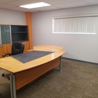 Empty office with a curved wooden desk, black chair, cabinet, and window blinds