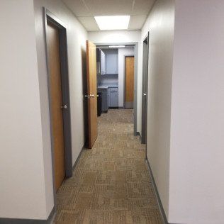 Narrow office hallway with beige carpet, white walls, and open wooden doors leading to a small kitchen area.