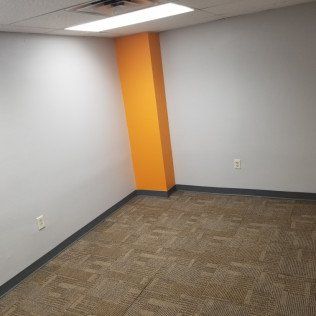 Empty office corner with gray walls, orange accent column, and carpeted floor.