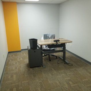 Empty office with a desk, black chair, trash bin, and yellow accent wall on carpeted floor