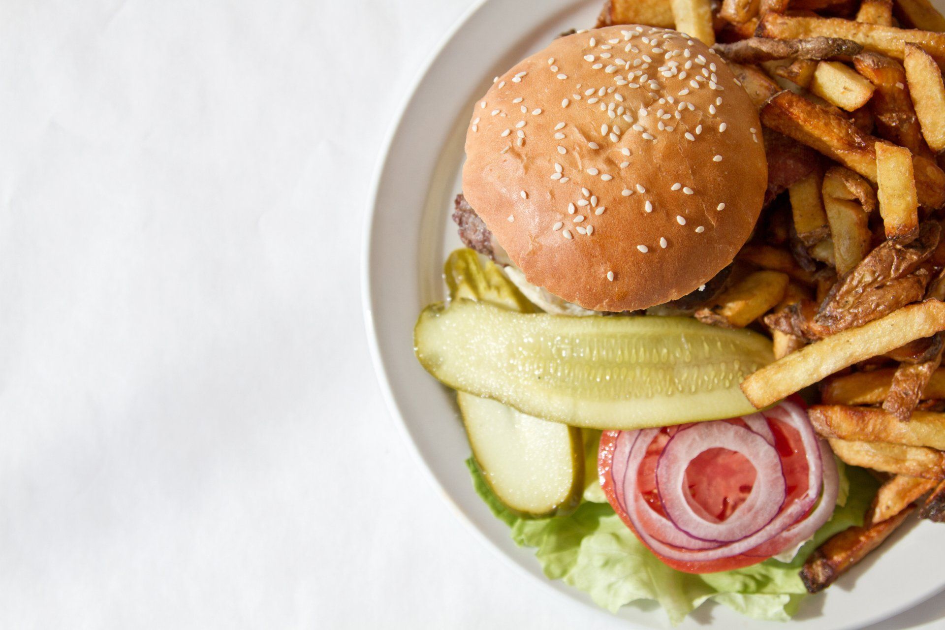 Hamburger on sesame bun with fries, pickle, red onion, and lettuce on white plate.