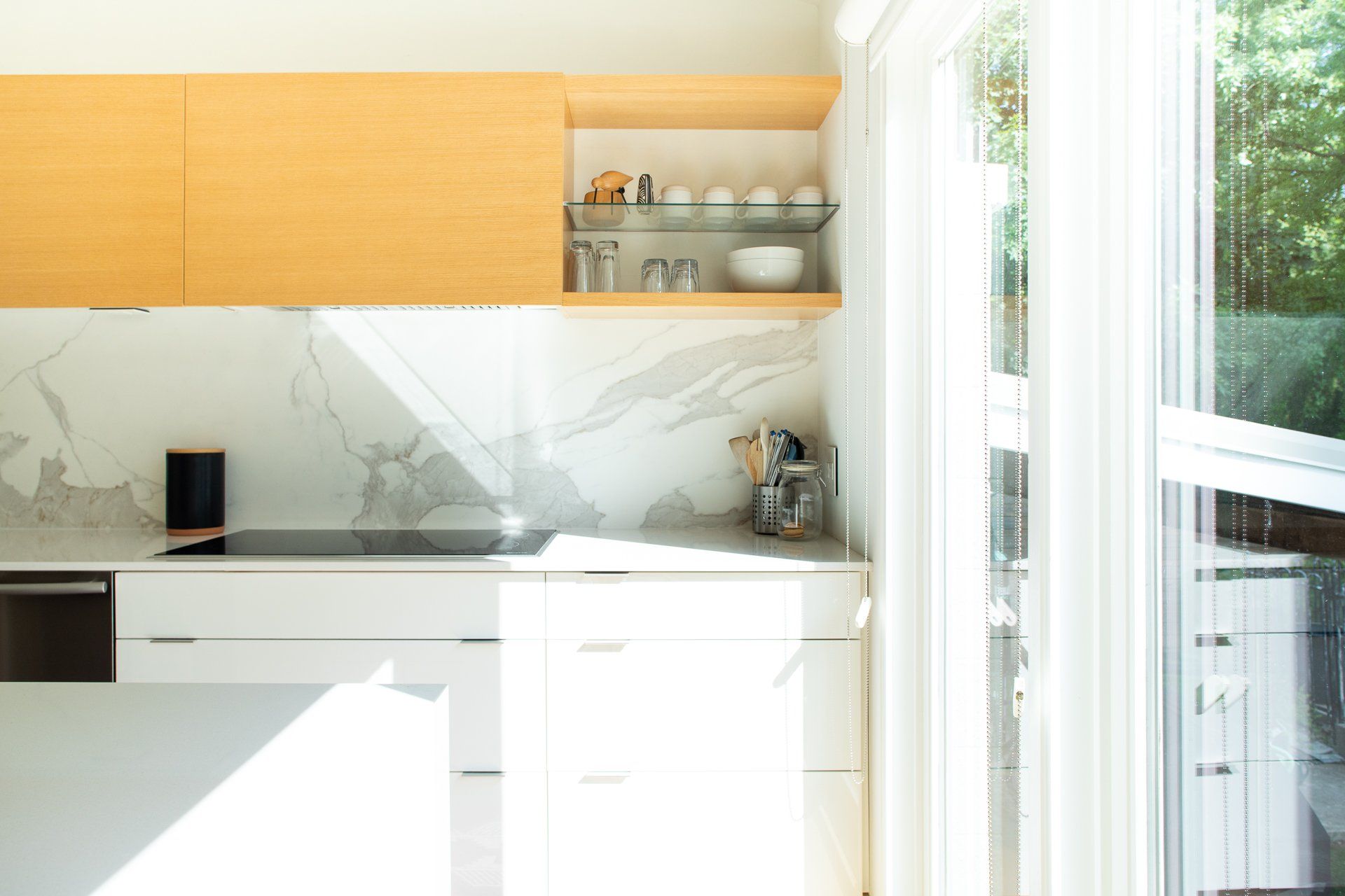 Modern kitchen with white cabinets, marble backsplash, and wooden accents, with sunlight streaming through a window.