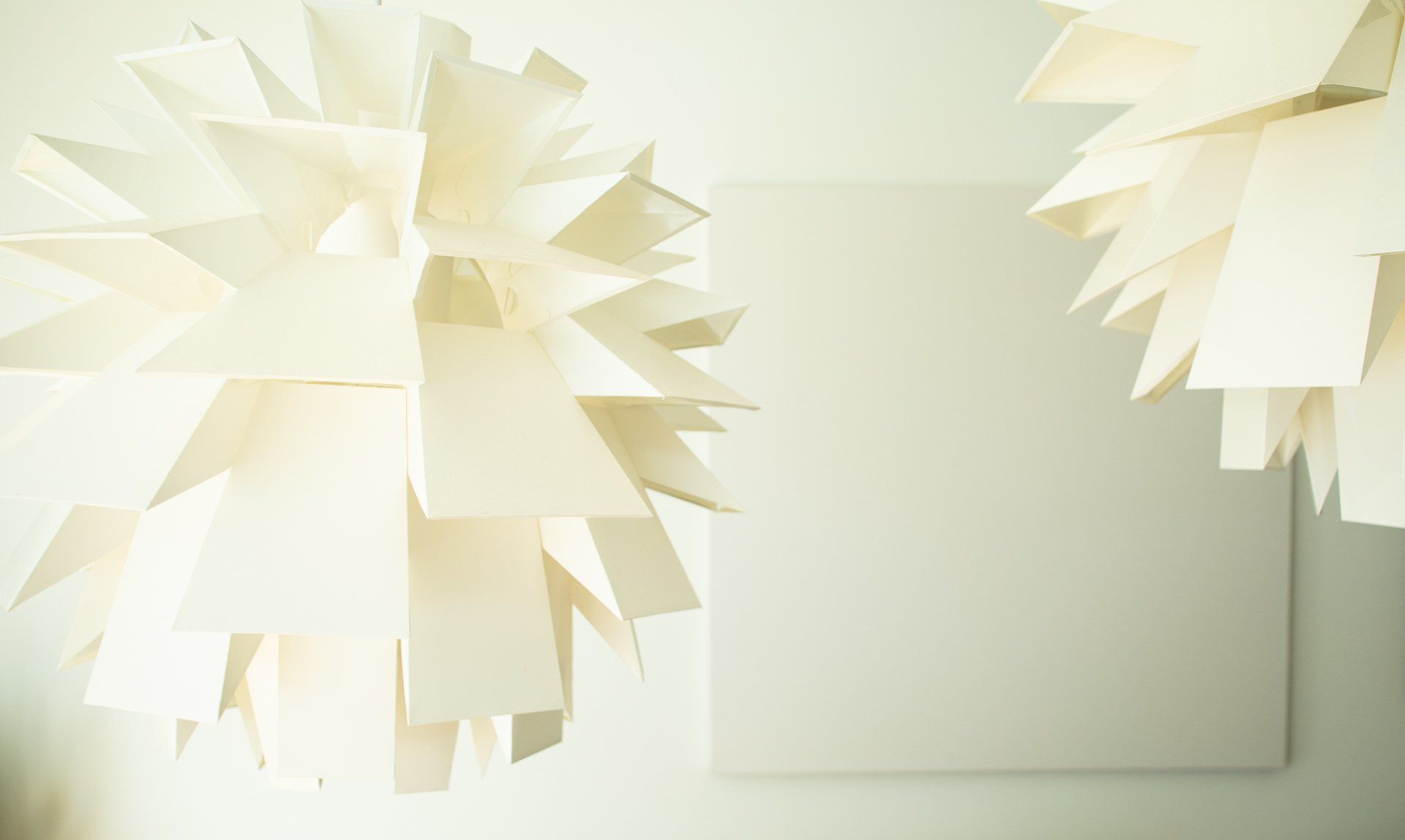 Two white origami-style pendant lamps flank a blank, square canvas on a light-colored wall.