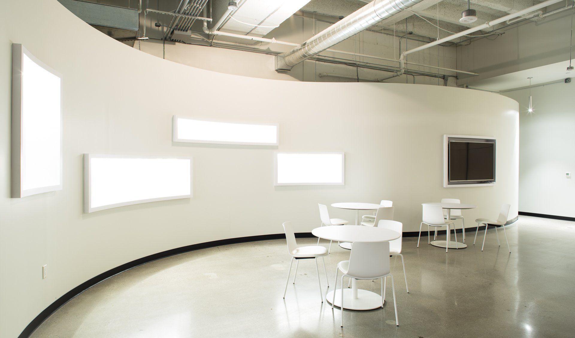 Modern, white-walled room with a curved wall featuring bright rectangular displays. White tables and chairs on polished concrete floor.