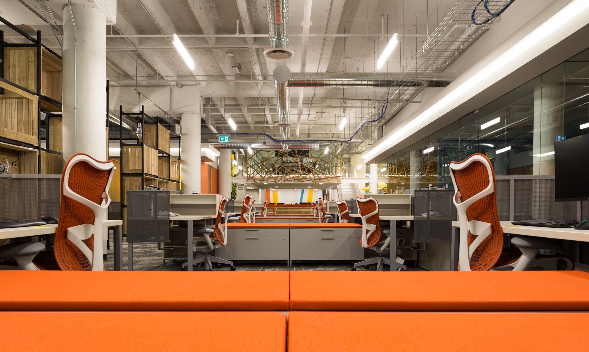Bright orange modern office with rows of desks, chairs, and benches. White support beams, and natural light.