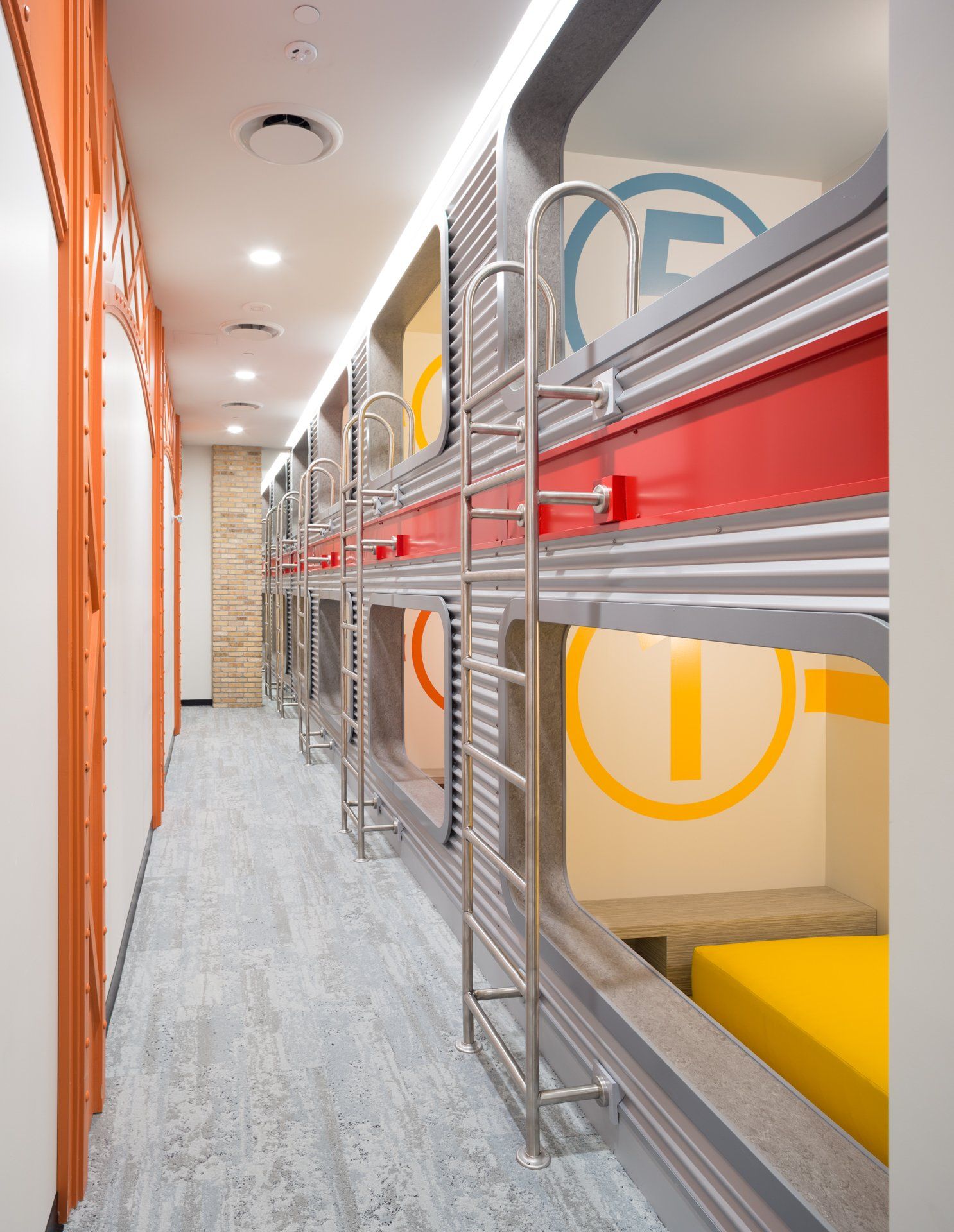 Capsule hotel hallway with stacked sleeping pods in red, yellow, and gray, with ladders.