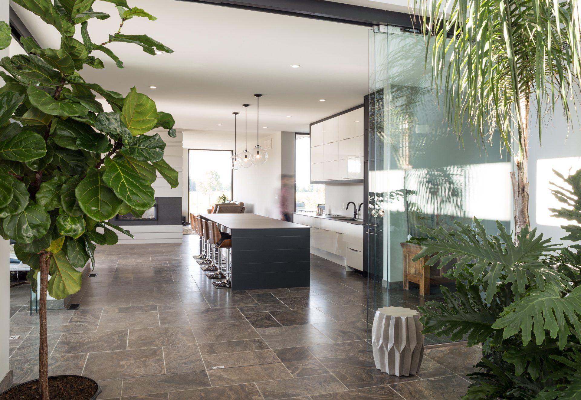 Modern open-plan kitchen with large island, plants, and natural light. Grey stone floor.