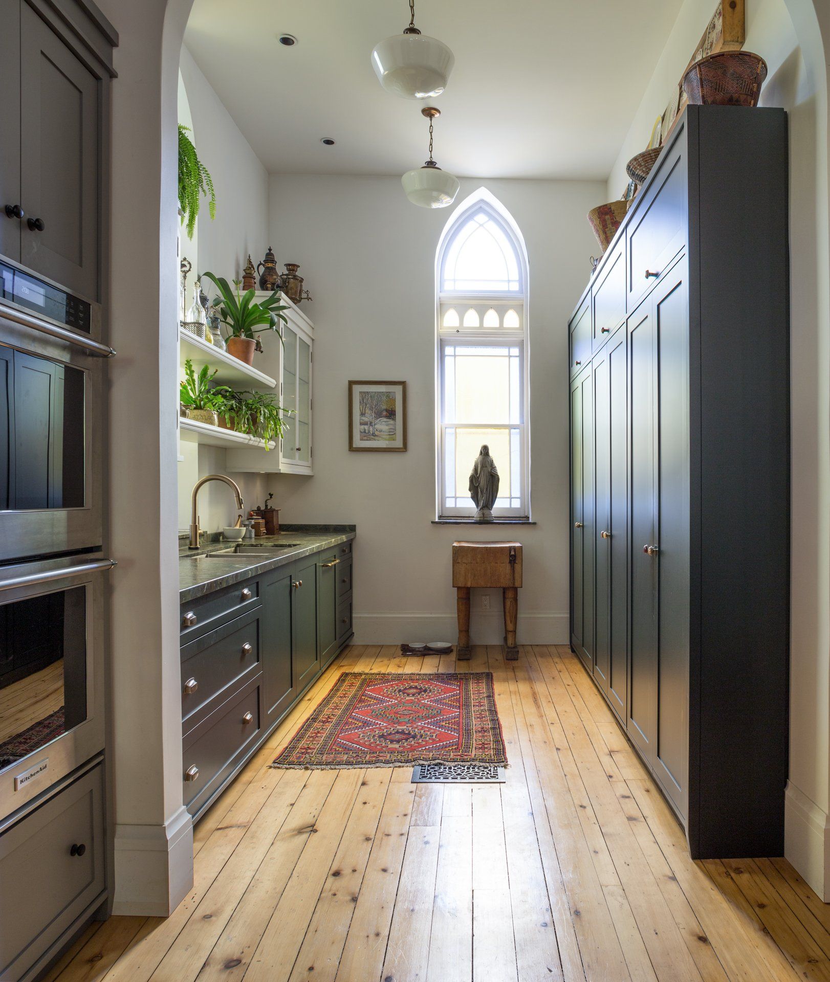 Narrow kitchen with dark green cabinets, wooden floors, and a gothic-arched window.