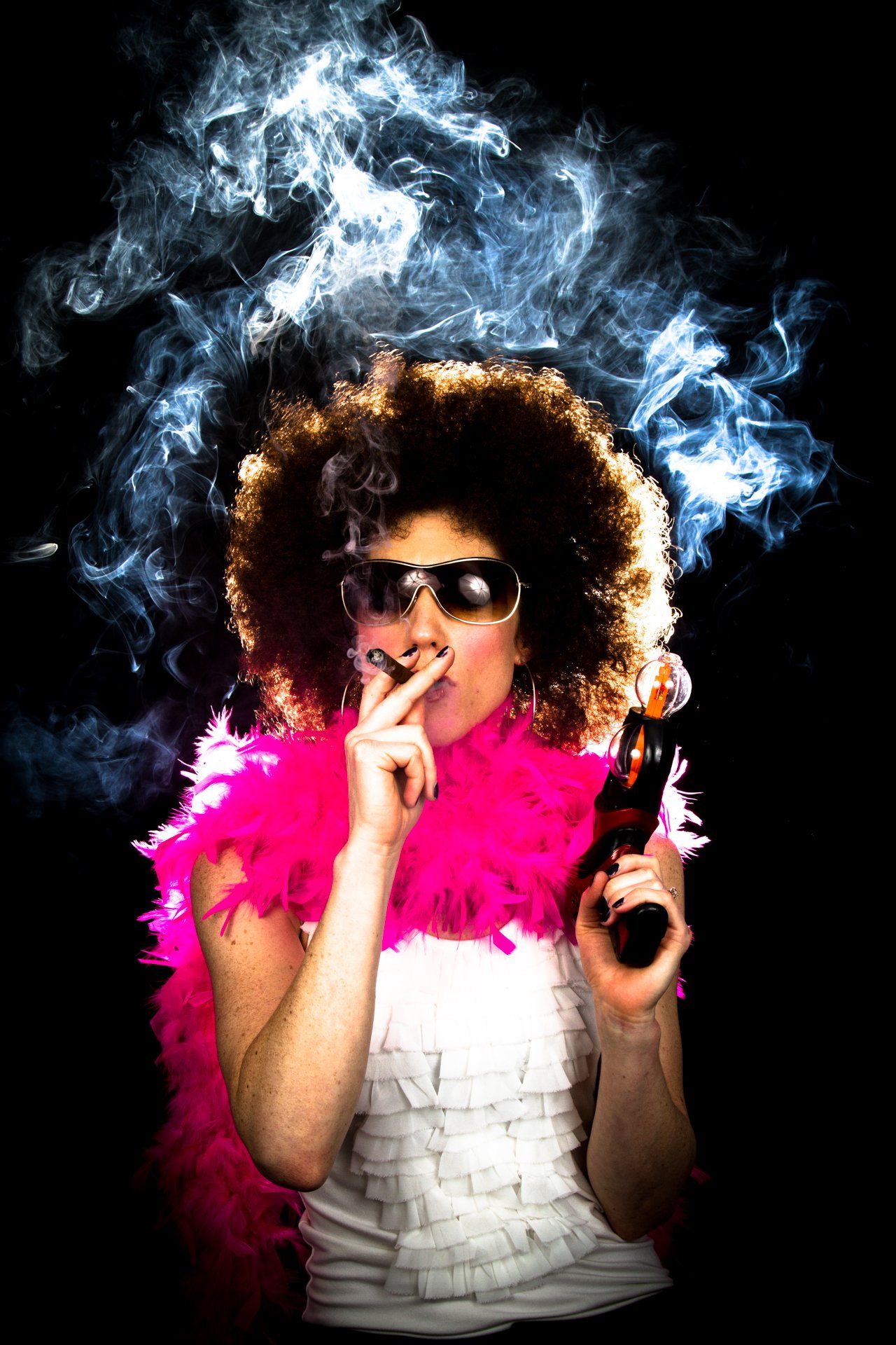 Woman in afro, sunglasses, and pink boa, smoking, holding a gun, with smoke cloud.
