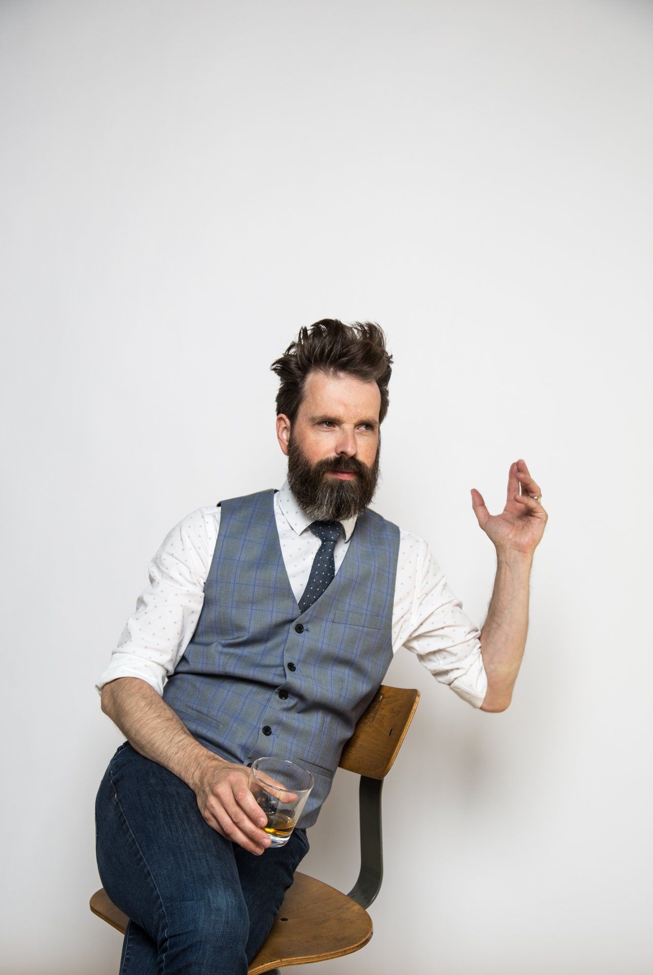 Man with beard in vest and tie, sitting on a wooden chair, holding a glass, and gesturing with his hand.