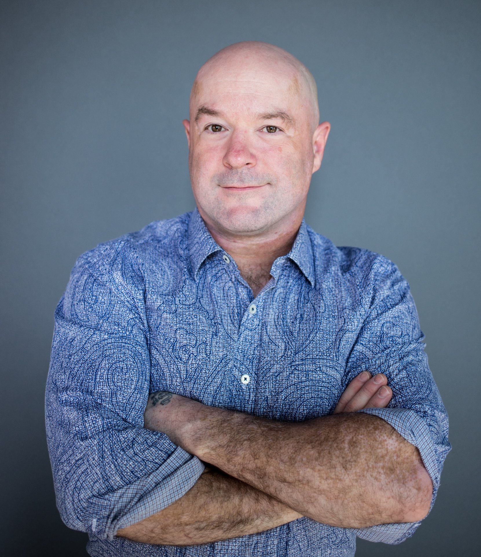 Bald man in blue patterned shirt with arms crossed, smiling. Gray background.
