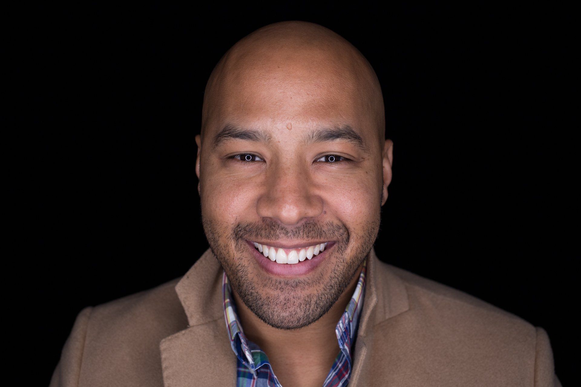 Smiling man with shaved head, wearing a tan blazer and plaid shirt, against a black background.