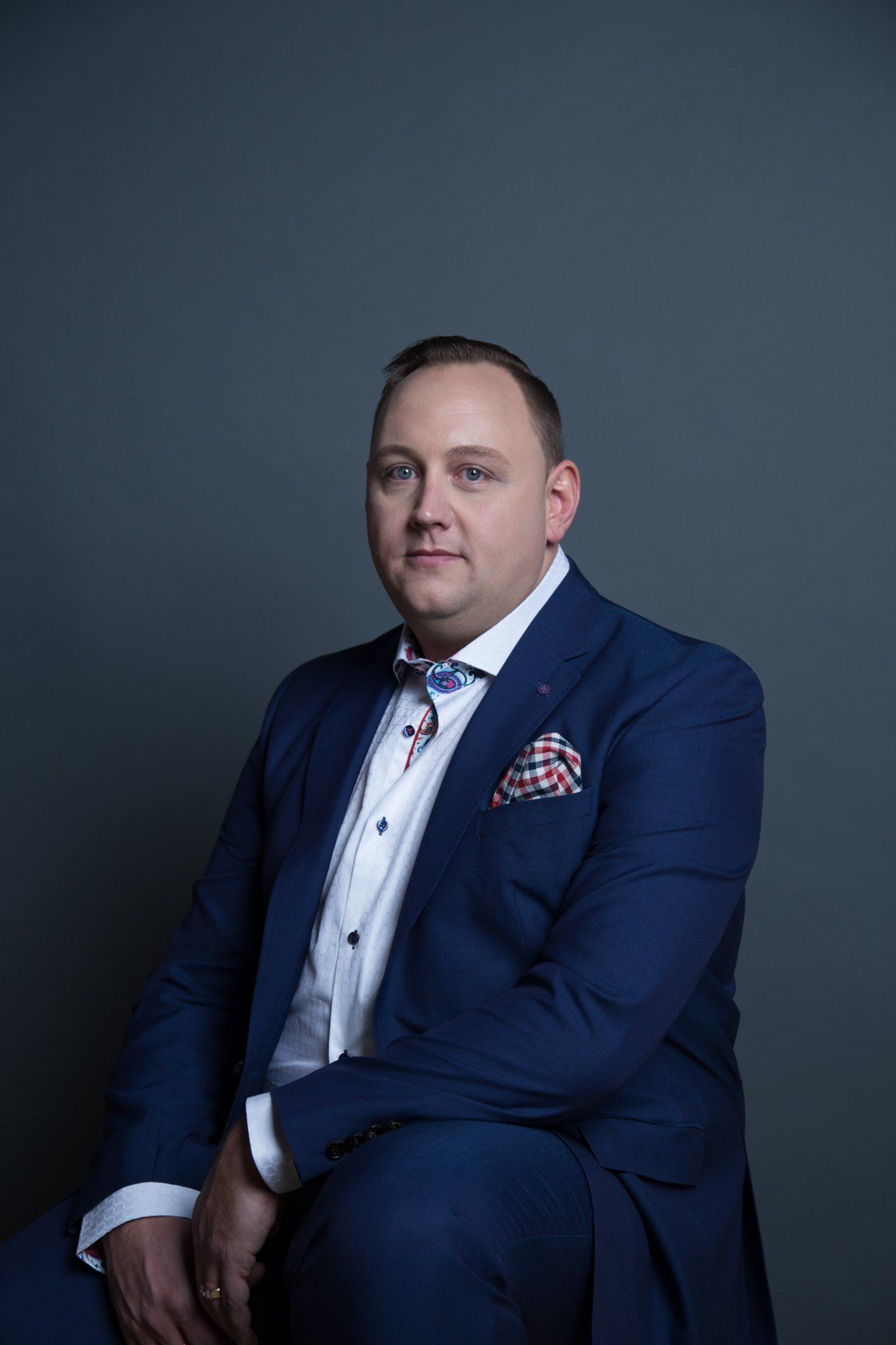 Man in a blue suit with a bow tie and pocket square, sitting against a gray background.