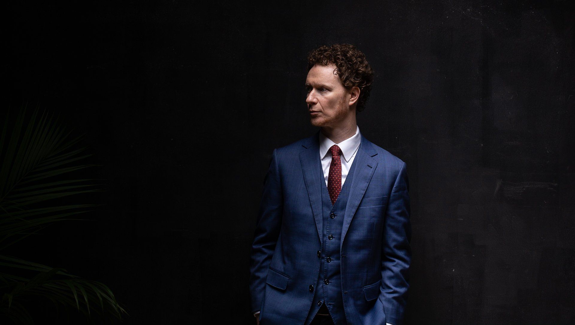 Man in blue suit, looking left, hands in pockets, against a dark background with a plant.