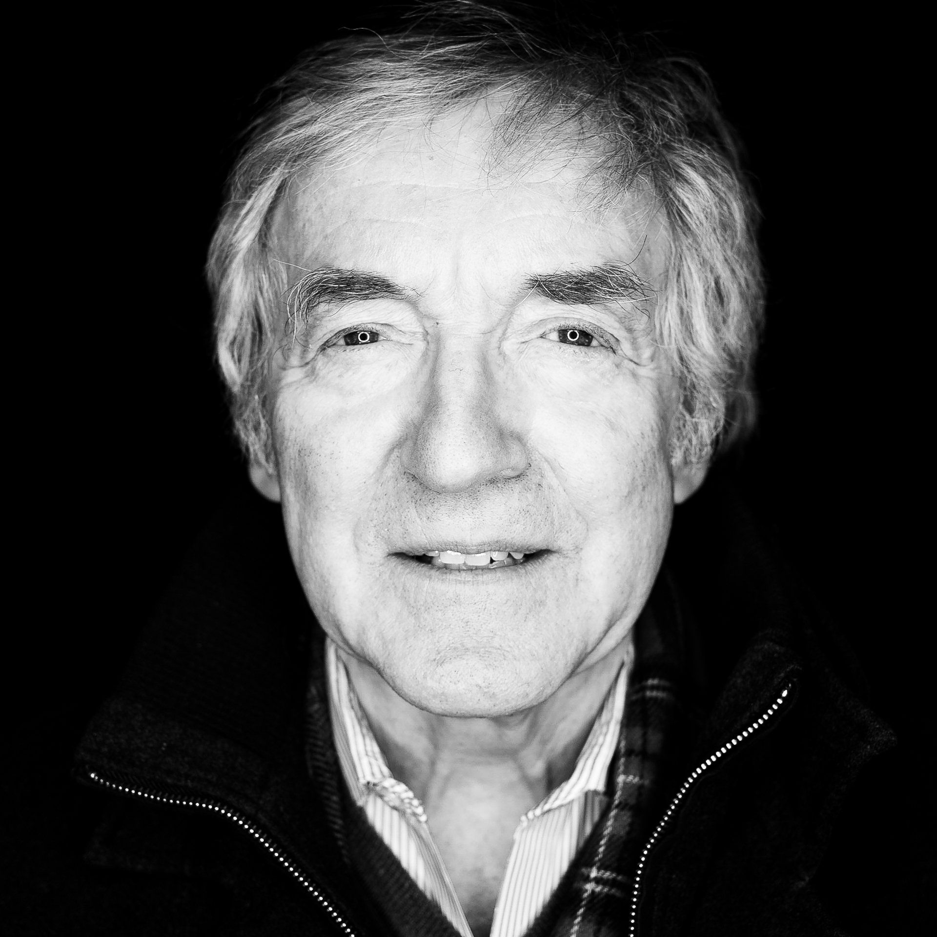 Man in black and white portrait, smiling slightly. Dark background, light hair, textured clothing.