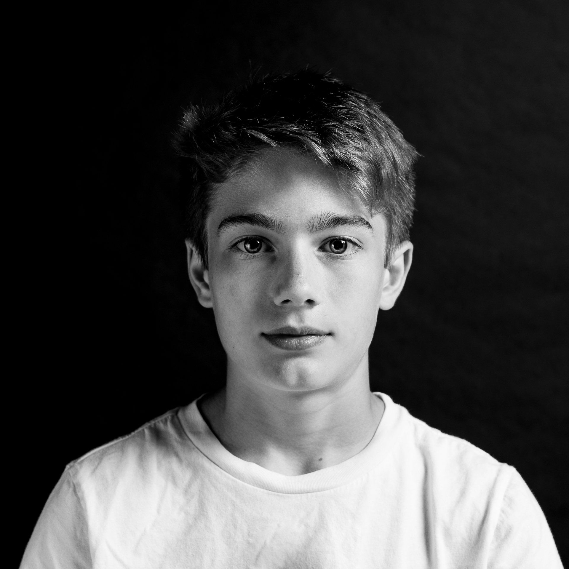 Teenager in white t-shirt against a dark backdrop, looking at the viewer.