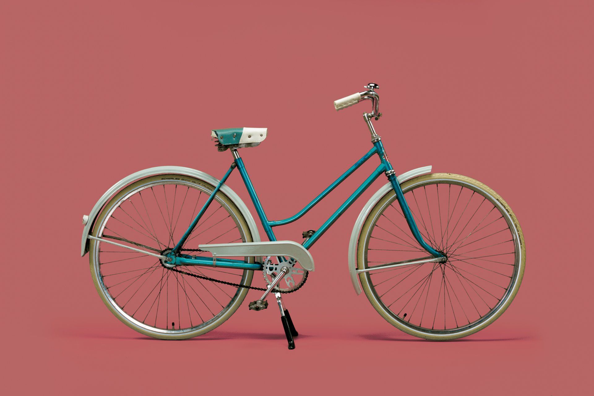 Teal vintage bicycle with white fenders and saddle against a pink background.