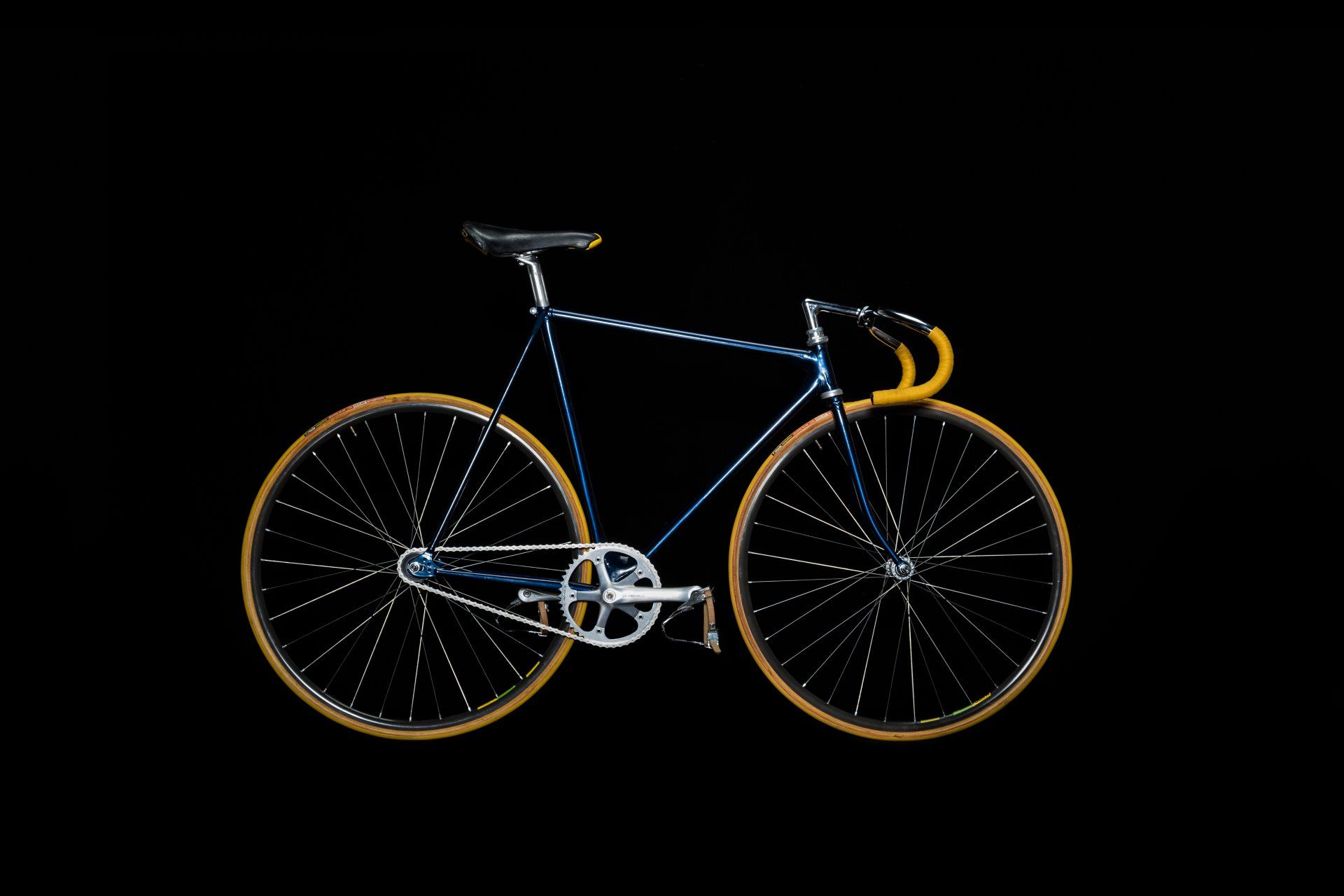 Blue and silver fixed-gear bicycle with tan tires and handlebars against a black background.