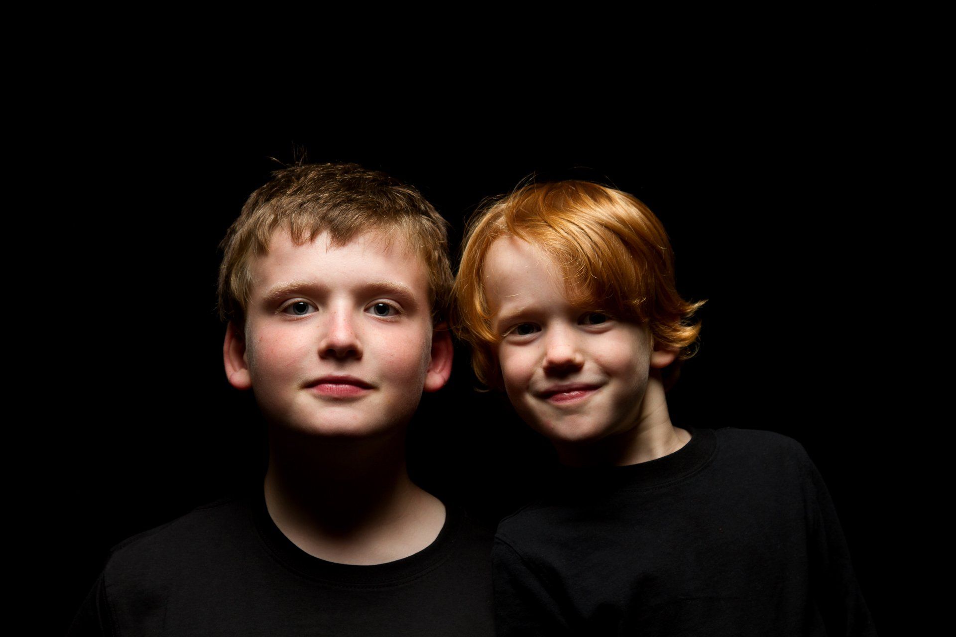 Two boys in dark shirts smiling against a black background. One with light brown hair, one with red hair.