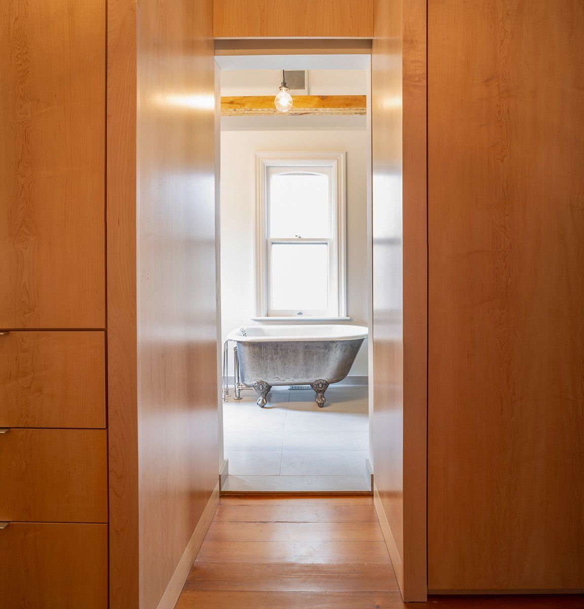 Hallway leading to a bathroom. Wooden cabinetry frames a view of a claw-foot tub near a window.