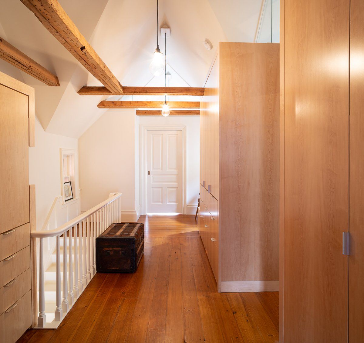 Hallway with wood floors, exposed beams, a trunk, and built-in cabinetry. White walls and door.
