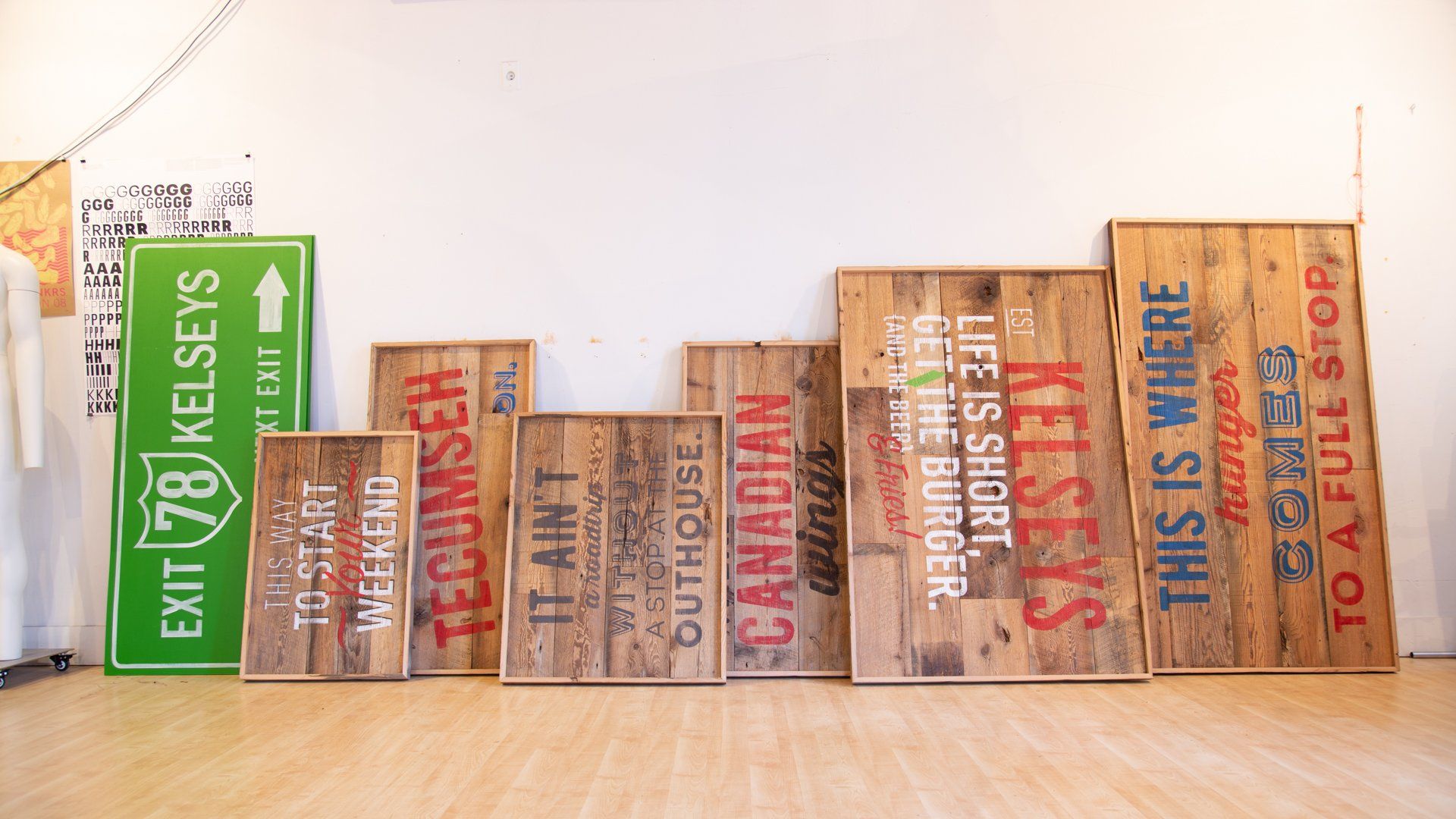 Collection of wooden signs with painted text and a green road sign against a white wall and wood floor.