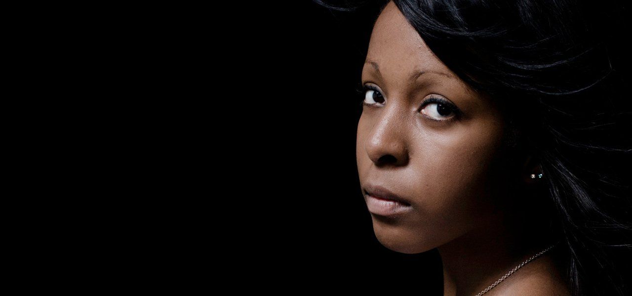 A person with dark skin looks toward the viewer, face centered in front of a dark background.