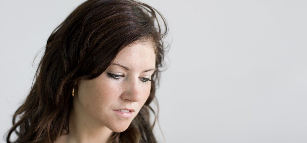 Woman with long, wavy brown hair looks down, set against a white background.