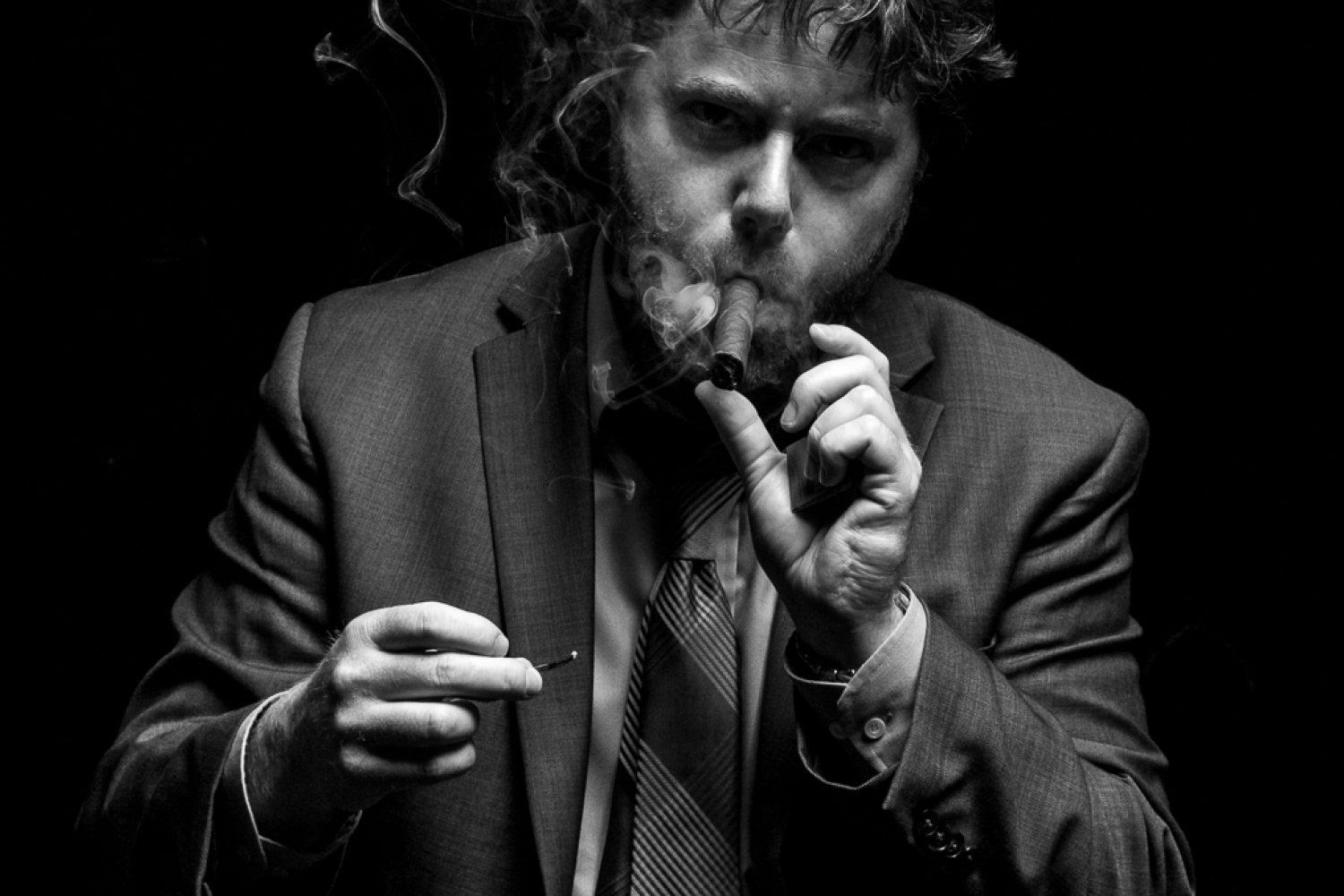Man in suit exhaling cigar smoke, dark background.