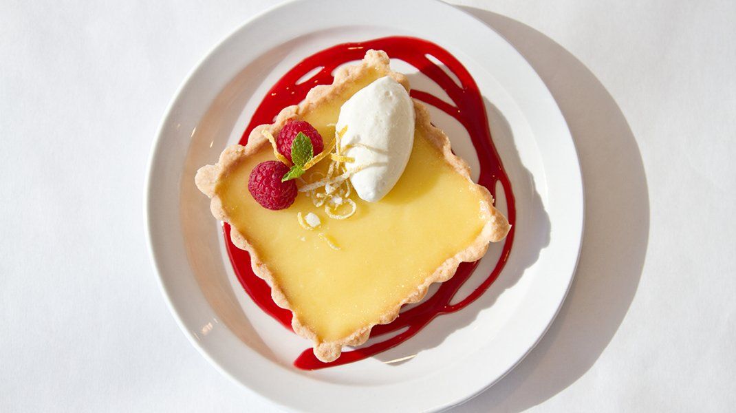 Lemon tart with raspberry sauce, whipped cream, and fresh raspberries on a white plate.