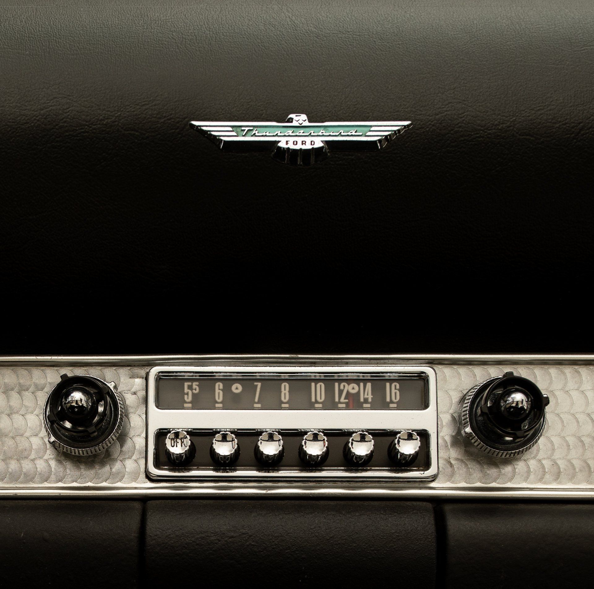 Vintage car dashboard with radio. Silver, chrome, black, and turquoise Thunderbird emblem.