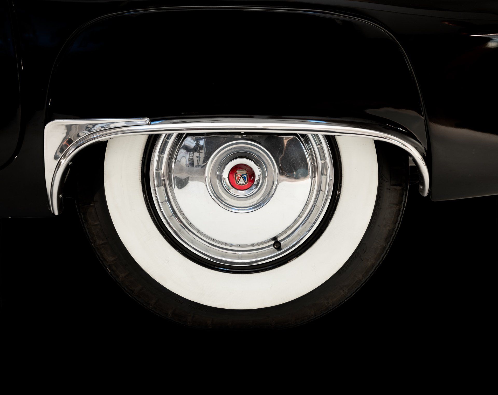 Close-up of a classic black car's rear wheel with white wall tire and chrome hubcap against a black background.