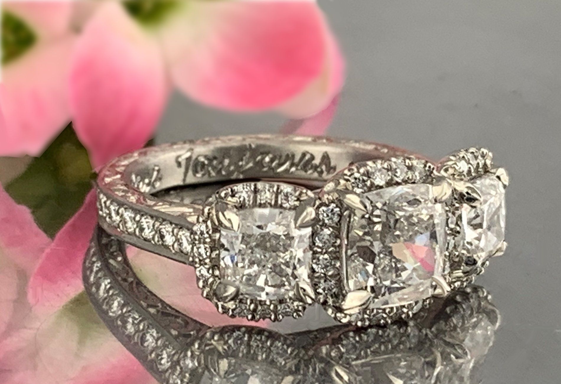 A diamond ring is sitting on a table next to a pink flower.
