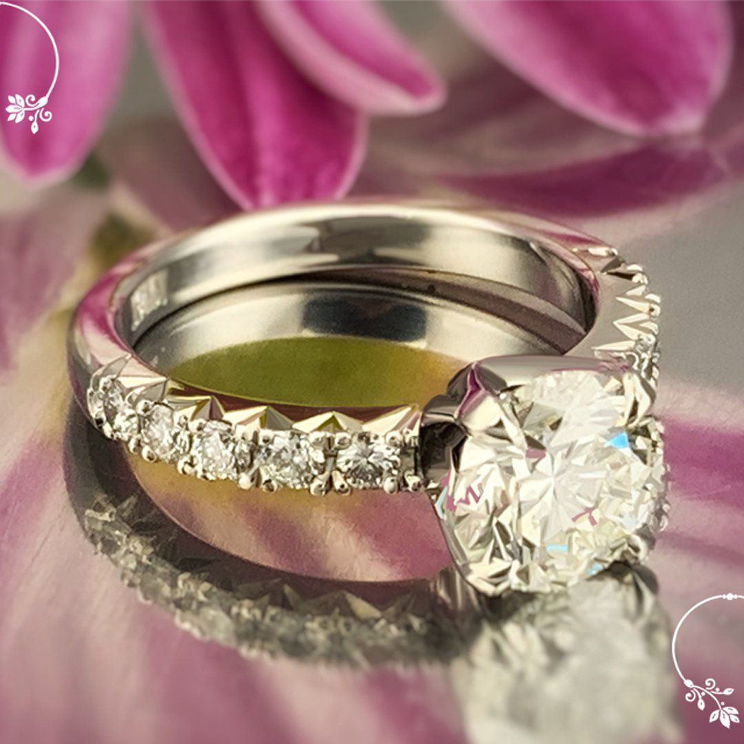 A wedding ring and engagement ring are on a table in front of a pink flower