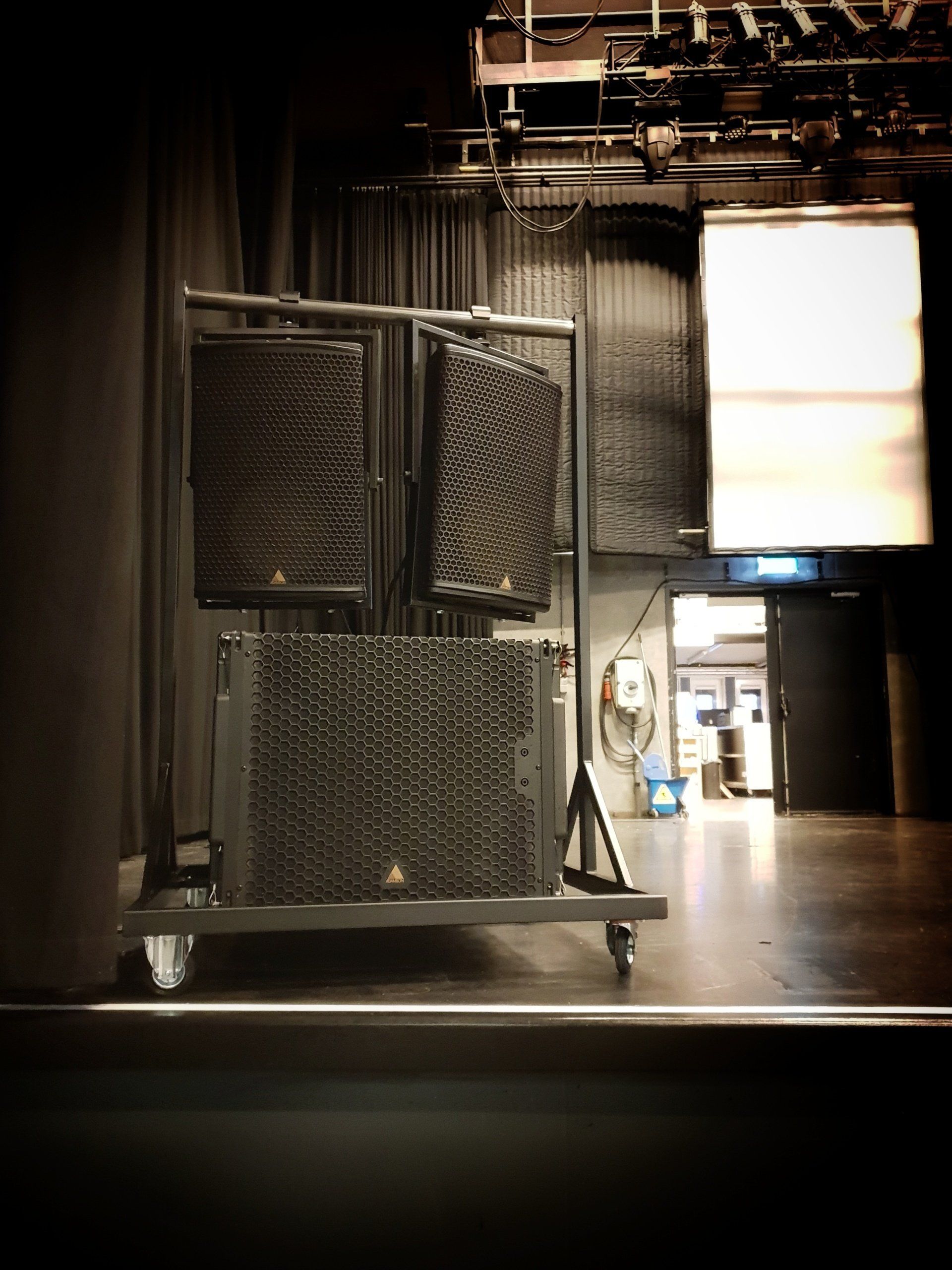 Op een donkere podiumvloer staat een verplaatsbaar metalen rek met drie zwarte, professionele luidsprekerkasten.