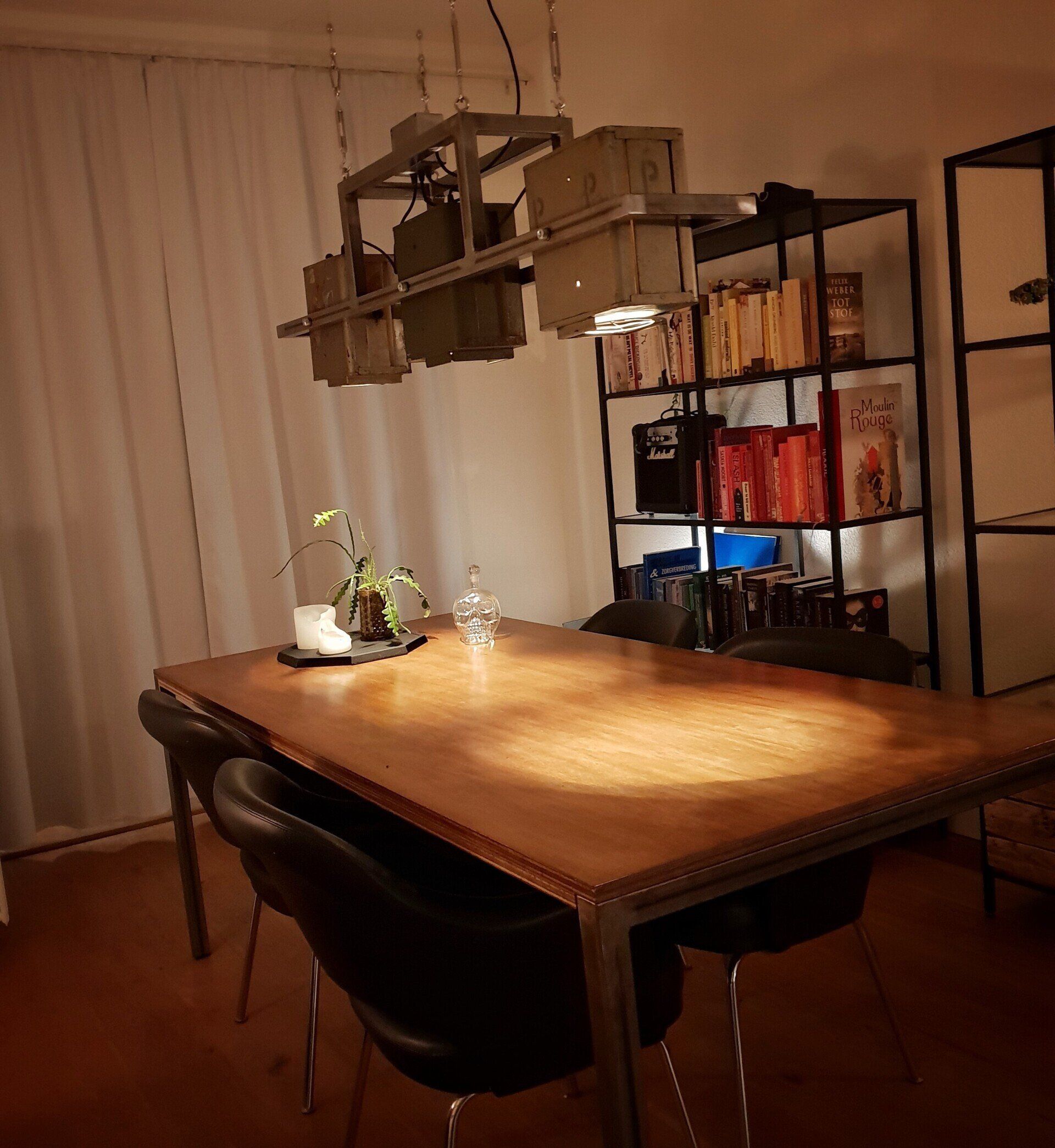 Een eetkamer met een houten tafel, zwarte stoelen, een unieke industriële lamp en een metalen boekenkast.
