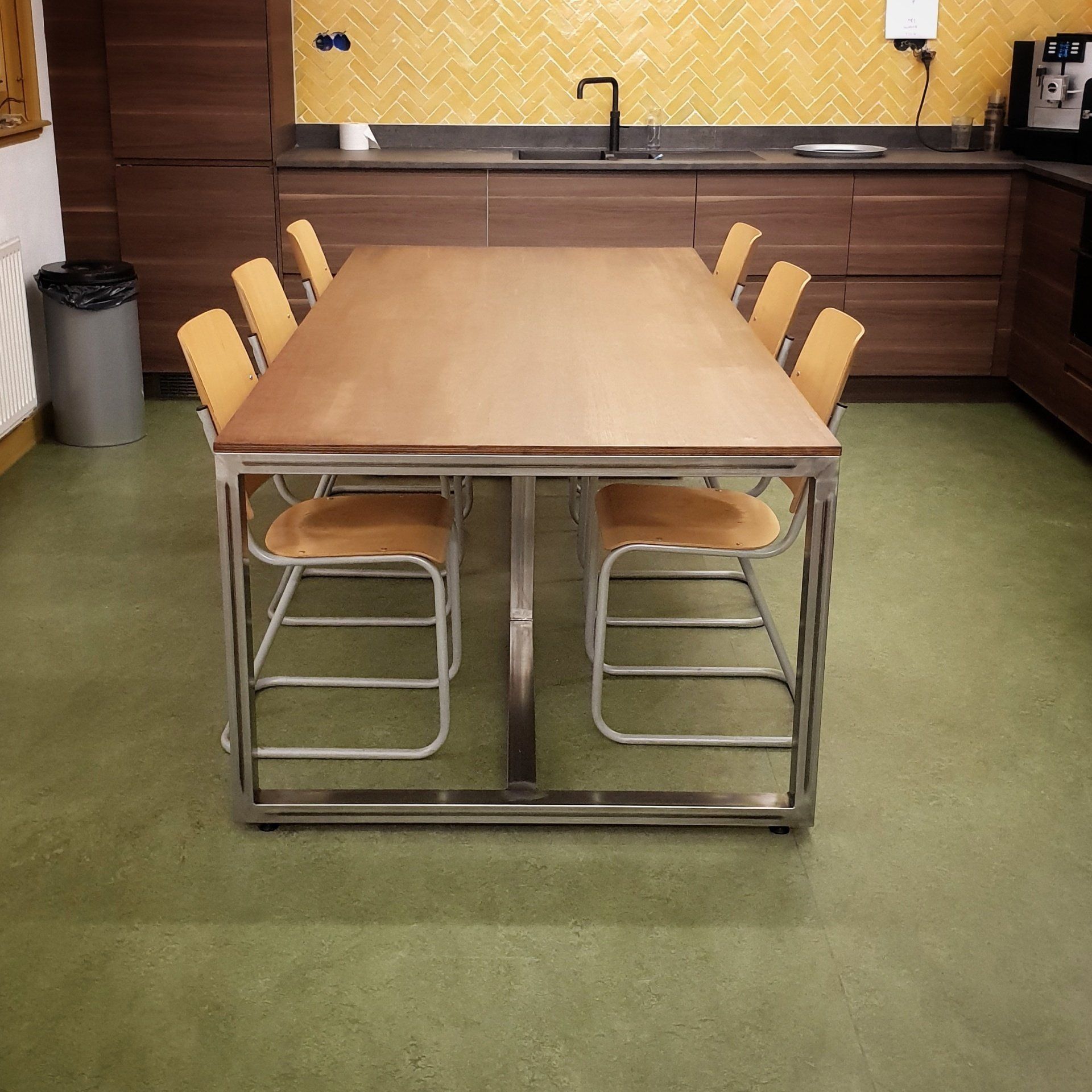 Een grote houten tafel met een metalen frame en bijpassende stoelen in een keuken met een gele tegelwand en bruine keukenkasten.