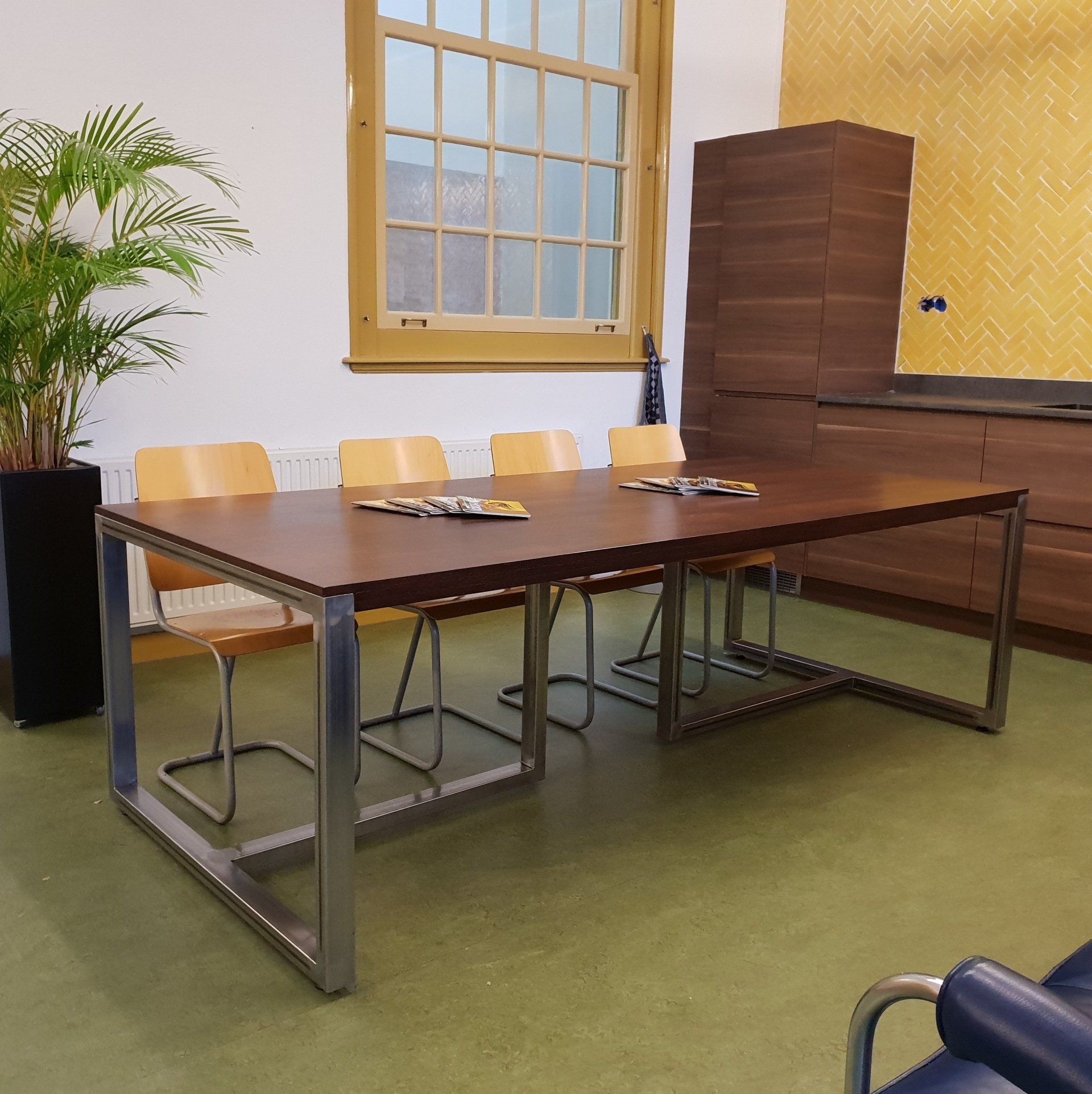 Een donkerhouten vergadertafel met metalen poten, vier lichthouten stoelen en een potplant in een lichte kantoorruimte.