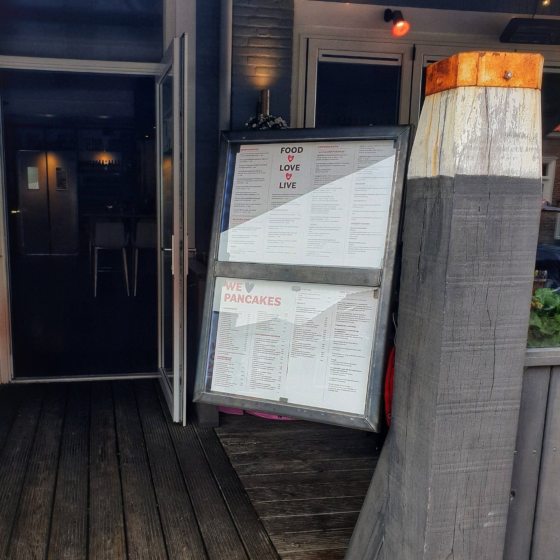 Een menubord staat op een houten terras buiten een deuropening, naast een grote, verweerde verticale houten paal.
