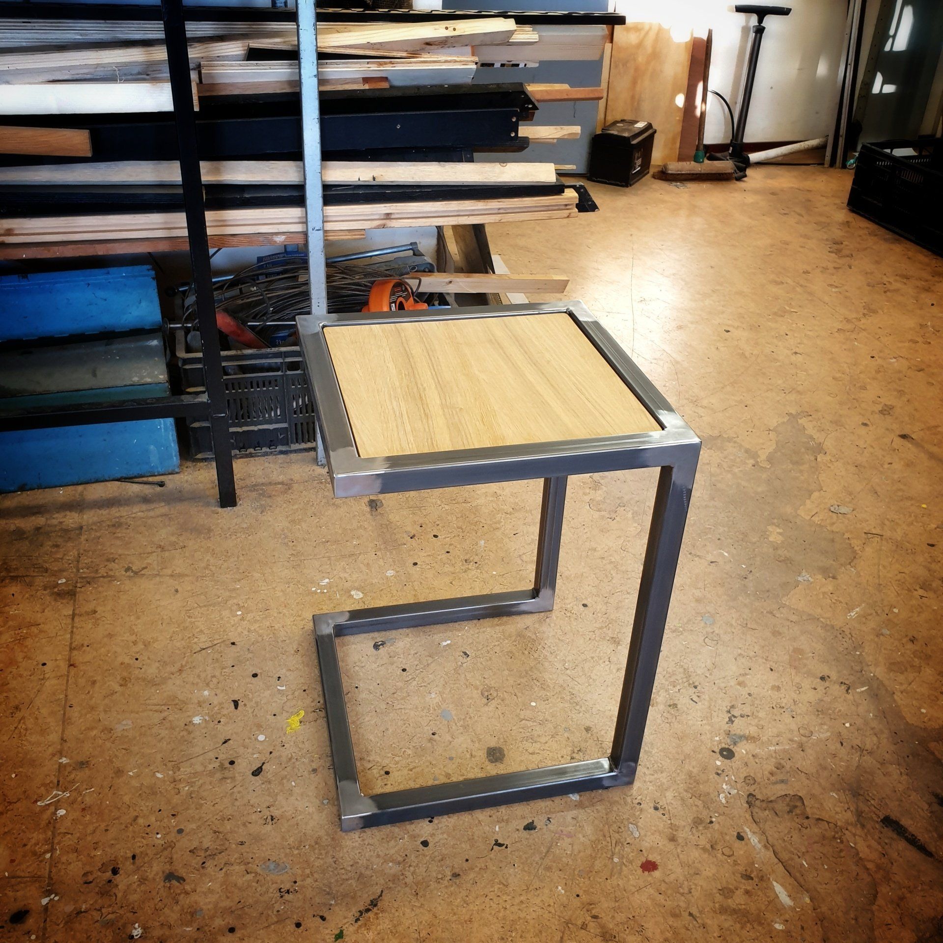 Een klein C-vormig bijzettafeltje met een vierkant houten blad en een grijs metalen frame, dat op de vloer van een werkplaats staat.
