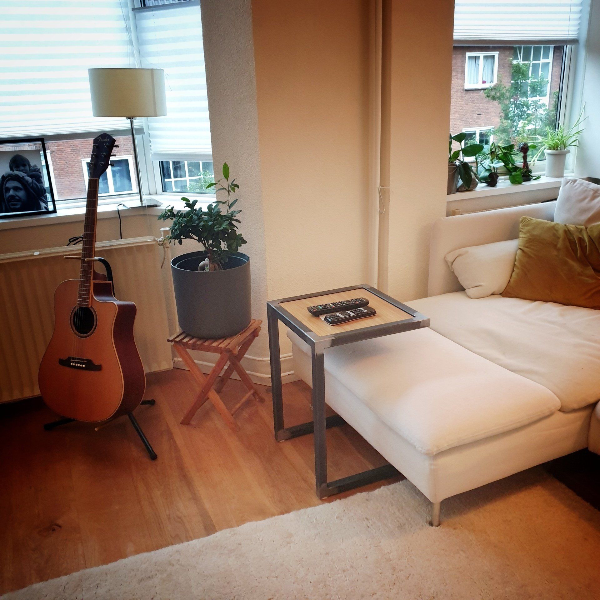 Een knusse woonkamer met een gitaar op een standaard, een potplant en een metalen C-vormig bijzettafeltje naast een witte bank.